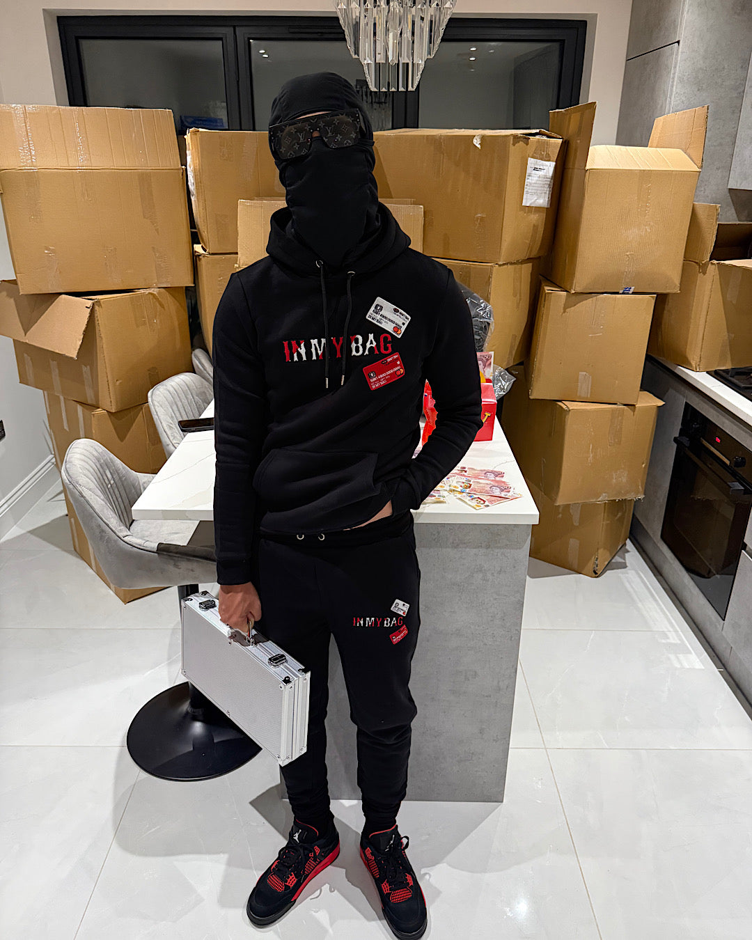 Person wearing a black hoodie and pants with visible branding, standing in a room with cardboard boxes.
