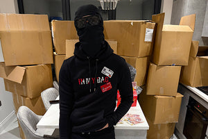 Person wearing a black hoodie and pants with visible branding, standing in a room with cardboard boxes.